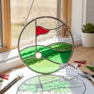 Puede incluir: Una escena de golf redonda de vidrieras con una bandera roja y hierba verde, colgada de una cadena. Una pequeña etiqueta de madera con la palabra "Dad" está adjunta. También se ven herramientas y materiales para crear vidrieras.