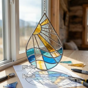 May include: A teardrop-shaped stained glass suncatcher with a sun and ocean design. The sun is yellow, and the ocean is blue and turquoise. The suncatcher hangs from a chain. A paper with a design and tools are on a wooden surface.