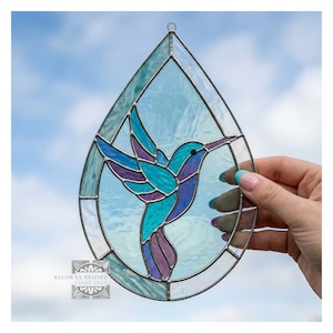 May include: A stained glass hummingbird in shades of blue, teal, and purple, set within a teardrop-shaped frame. The artwork features a detailed design and is held against a backdrop of a blue sky.