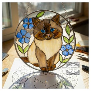 Op de afbeelding: Rond gebrandschilderd glazen kunstwerk met een kat met blauwe ogen, blauwe bloemen en groene bladeren. De kat heeft bruine en crèmekleurige vacht. Het stuk hangt aan een ketting. De tekst "KELOWNA STAINED GLASS SINCE 2023" is zichtbaar.