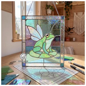 Pode incluir: Um painel de vitral com um sapo verde com asas de fada, sentado numa folha de nenúfar. O painel quadrado, com borda transparente, é suspenso por uma corrente. O sapo é cercado por vidro iridescente em tons de verde, azul e roxo.