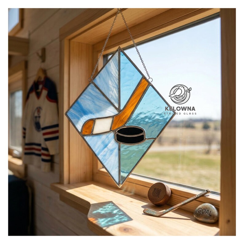 May include: Stained glass hockey-themed window art. The diamond-shaped piece features a hockey stick, puck, and blue and white glass. The piece is hanging in a wooden window frame. The logo "KELOWNA STAINED GLASS" is visible.