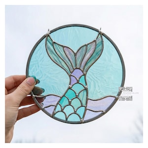 May include: A stained glass mermaid's tail in teal, purple, and blue, set within a circular frame. The glass has a textured appearance, and the design is held together with dark metal. The artwork is held by a hand.
