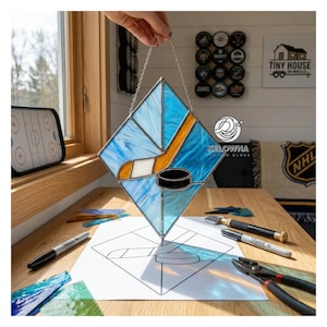 May include: A stained glass hockey-themed piece in a diamond shape, featuring a hockey stick, puck, and blue and white glass. The piece is suspended by a chain. The word "Kelowna" is visible.