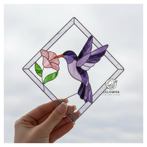 May include: Stained glass hummingbird art piece. The design features a purple hummingbird with outstretched wings, feeding from a pink flower. The artwork is set within a diamond-shaped frame. The logo "KELOWNA STAINED GLASS" is visible.