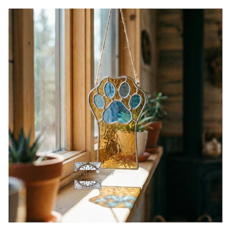 May include: Stained glass paw print suncatcher with blue and gold hues, hanging in front of a window. The paw print design is outlined in black and suspended by a natural fiber cord. A decorative item for pet lovers.