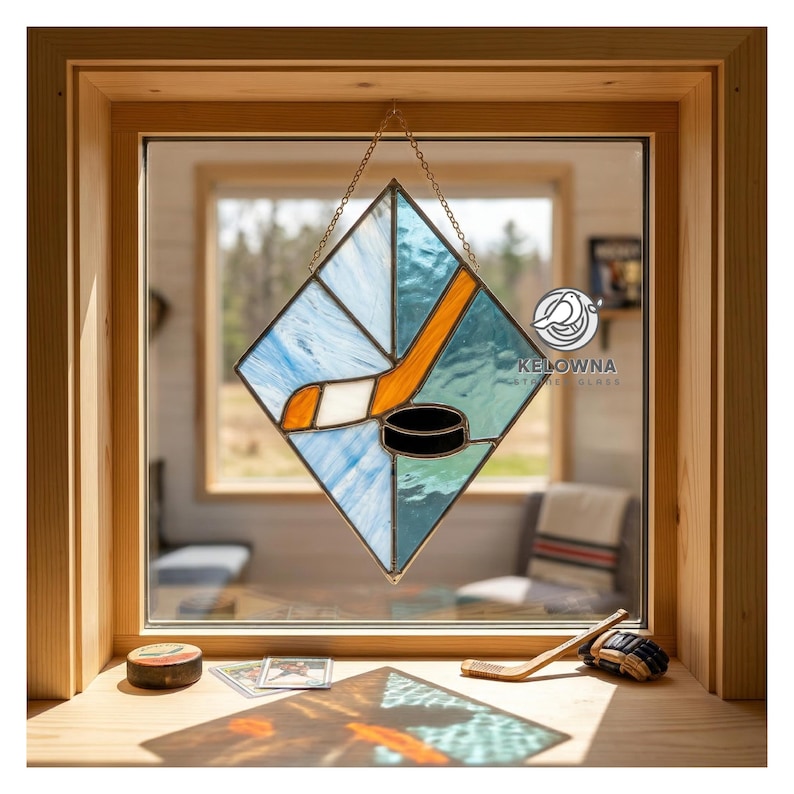 May include: Stained glass hockey-themed window art. The diamond-shaped piece features a hockey stick, puck, and blue and white glass. The piece is suspended by a chain. The window frame is made of wood.