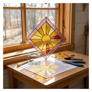 Pode incluir: Um apanhador de sol em vitral em forma de losango, com um desenho de sol em tons de amarelo, laranja e vermelho. Pendura-se por uma corrente. Um modelo de design e ferramentas estão sobre a mesa. Dimensões: cerca de 20 cm x 20 cm.