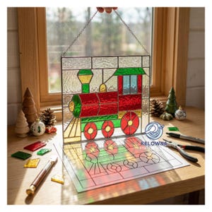 Puede incluir: Un panel de vidrieras que representa un tren rojo, verde y amarillo, suspendido por una cadena. El diseño del tren está sobre un fondo de vidrio transparente. El panel se exhibe cerca de una ventana con decoraciones navideñas.