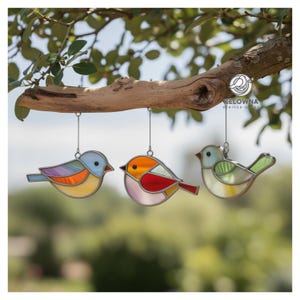 Peut inclure: Trois ornements d'oiseaux en vitrail colorés suspendus à une branche d'arbre. Les oiseaux présentent des couleurs vives, notamment le bleu, l'orange, le rouge et le vert. Chaque oiseau est suspendu par un fil fin, créant un affichage décoratif.