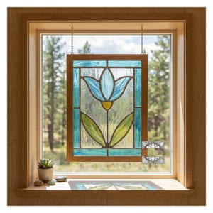 May include: Stained glass window panel featuring a blue tulip design with green leaves and a yellow center. The panel is framed in wood and hangs in front of a window with a view of trees.