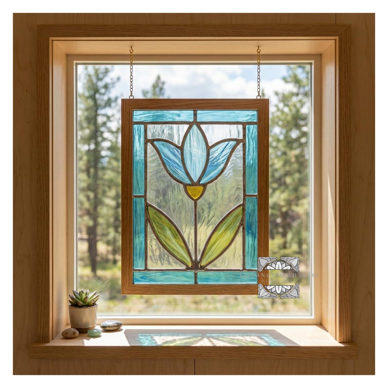 May include: Stained glass window panel featuring a blue tulip design with green leaves and a yellow center. The panel is framed in wood and hangs in front of a window with a view of trees.