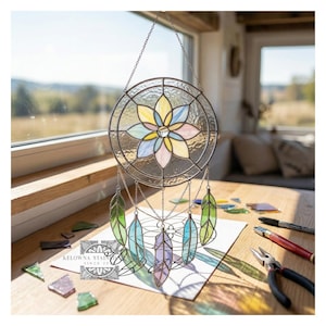 Op de afbeelding: Een gebrandschilderde dromenvanger met een bloemmotief in het midden, in pastelkleuren. Onder de cirkel hangen glazen veren in groen, blauw en paars. De dromenvanger wordt opgehangen aan een ketting.