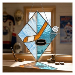 May include: Stained glass hockey-themed artwork. The piece features a hockey stick, puck, and ice rink design in shades of blue, orange, and black. The artwork is diamond-shaped and hangs from a chain. Additional hockey-related items are in the background.