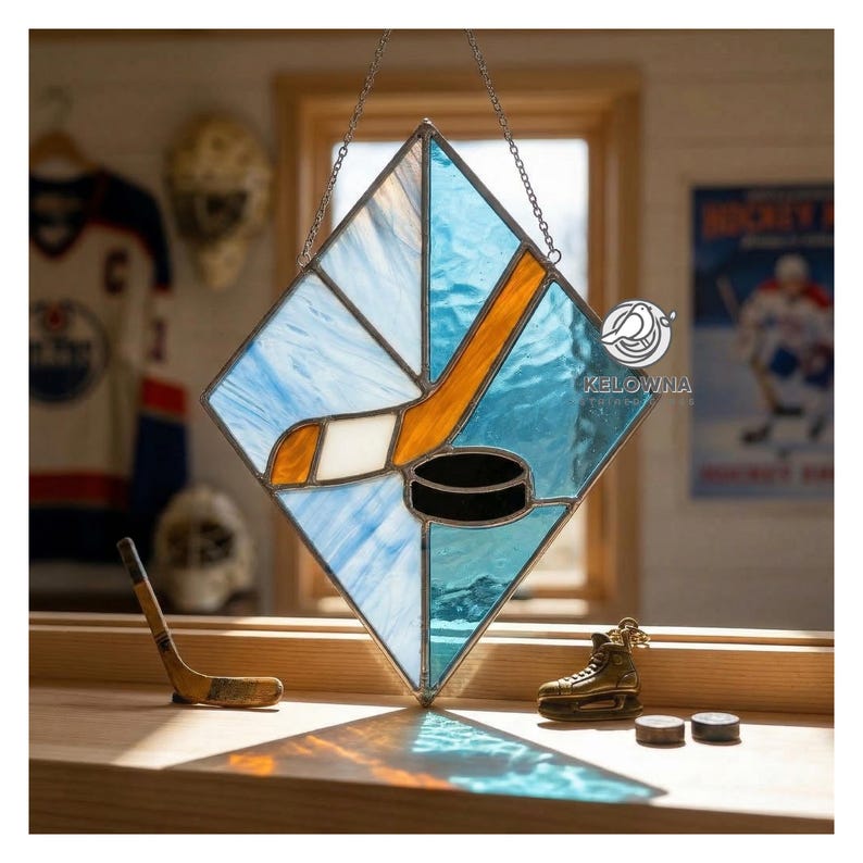 May include: Stained glass hockey-themed artwork. The piece features a hockey stick, puck, and ice rink design in shades of blue, orange, and black. The artwork is diamond-shaped and hangs from a chain. Additional hockey-related items are in the background.