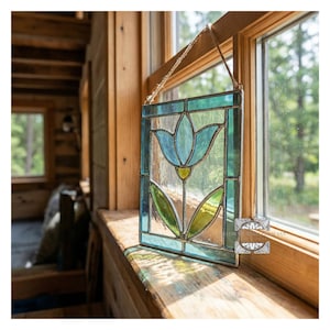 May include: A stained glass panel featuring a blue tulip design with green leaves and a yellow center. The panel is framed in teal and hangs from a natural fiber cord. The artwork is displayed in a wooden window frame.