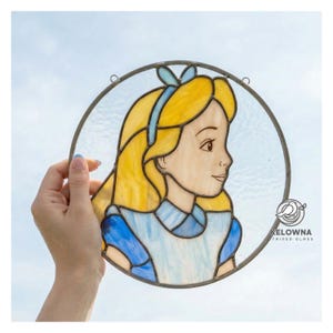 Peut inclure: Vitrail circulaire représentant Alice au pays des merveilles. L'œuvre d'art a un cadre en métal. Alice est représentée de profil avec des cheveux blonds, un bandeau bleu et une robe bleue. L'œuvre est présentée sur un fond de ciel bleu.