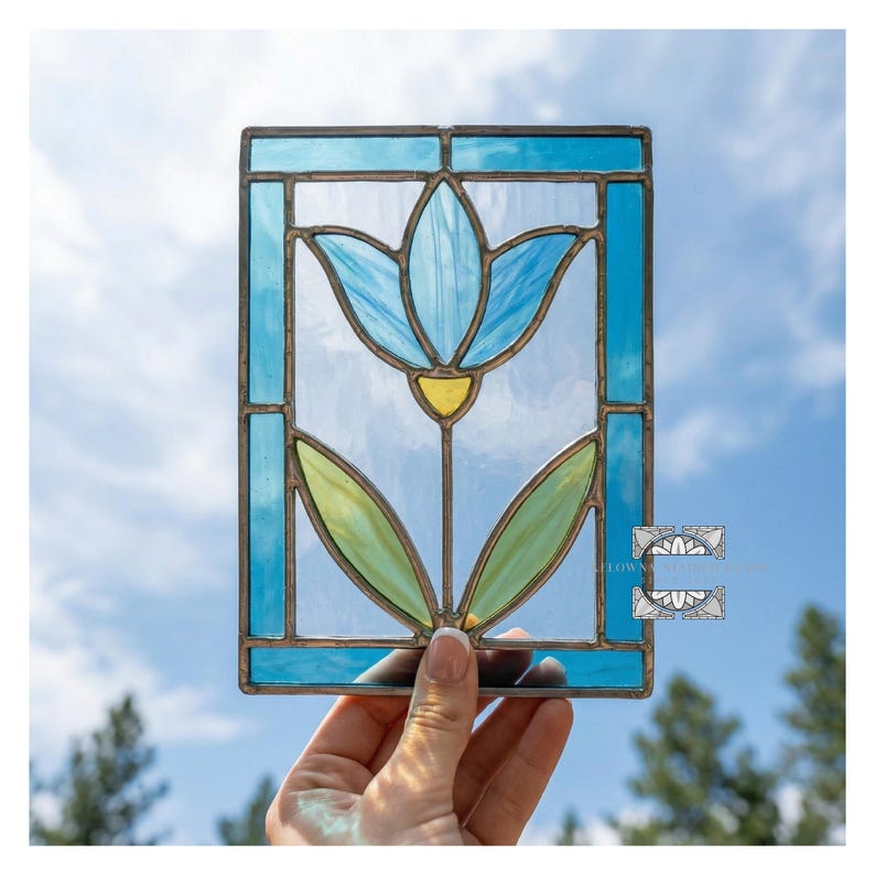 May include: A stained glass panel featuring a blue tulip flower with yellow accents, framed by a turquoise border. The flower has green leaves. The panel is held up against a blue sky.