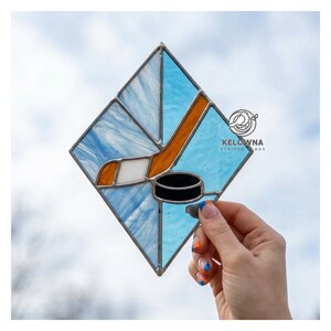 May include: Stained glass hockey-themed artwork in a diamond shape. The design features a hockey stick, puck, and ice, with blue, orange, white, and black colors. The artwork is held up against a cloudy sky.