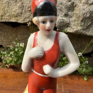 May include: A vintage porcelain figurine of a woman in a red swimsuit and a black and red swim cap. The figurine has a painted face with red lips and is posed with her arms crossed. The base is a light green color.