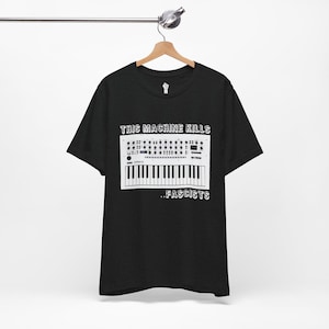 May include: Black t-shirt with a white graphic of a synthesizer and the text "THIS MACHINE KILLS ..FASCISTS". The shirt is hanging on a wooden hanger.