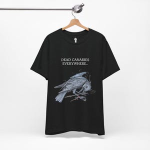 May include: Black t-shirt with the text "DEAD CANARIES EVERYWHERE.." above a detailed illustration of a crow holding a skull. The crow is perched on a branch, with intricate shading and detail. The shirt is hanging on a wooden hanger.
