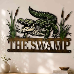 May include: A metal wall art piece featuring a green alligator with an open mouth, surrounded by cattails and tall grass. Below the alligator, a horizontal metal bar displays the text THE SWAMP in bold, capital, serif lettering.