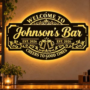 Peut inclure: Une enseigne murale en métal noir avec un rétroéclairage chaud affichant le texte "WELCOME TO Johnson's Bar EST. 2026 CHEERS TO GOOD TIMES" et un graphique de deux chopes de bière avec des ornements décoratifs sur un mur texturé.