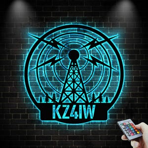 Könnte beinhalten: Ein schwarzes Metall-Wandkunstwerk mit einem Funkturm-Design und blauem Neon-Glow. Das Design umfasst eine kreisförmige Antenne, Blitze und den Text "KZ4IW". Die Kunst ist an einer dunklen Backsteinmauer angebracht.