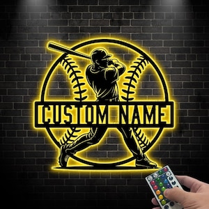 May include: A black and yellow baseball-themed sign with the text "CUSTOM NAME". The sign features a baseball player swinging a bat inside a baseball shape. The sign is illuminated with yellow light and is mounted on a dark brick wall.