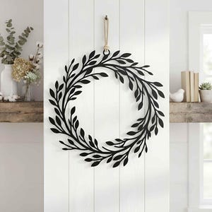 May include: A circular wall hanging featuring a matte black metal silhouette of a leafy wreath. The design consists of interconnected branches and oval-shaped leaves arranged in a continuous ring, suspended by a thick, light-coloured jute rope loop.