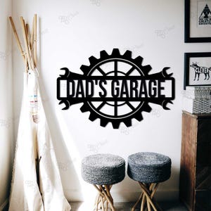 May include: Black metal wall art with the text "DAD'S GARAGE" inside a gear and wrench design. The sign is mounted on a white wall, with decorative elements in the background.