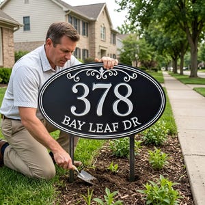 Puede incluir: Un letrero de dirección ovalado negro con números y texto blancos que dice "378 BAY LEAF DR" se está instalando en un jardín. El letrero está montado en dos postes de metal. La persona está usando una pequeña paleta para plantar el letrero.