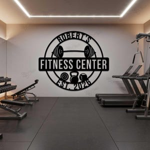 Può includere: Decorazione murale in metallo nero con la scritta "Robert's Fitness Center" e l'anno di fondazione "Est. 2026". Il design include un bilanciere, kettlebell e una cornice circolare, adatta per una palestra.