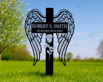 May include: Black metal memorial cross with angel wings and a rose design. The cross features the name "ROBERT S. SMITH" and dates 09.20.1962-06.15.2025. The cross is placed in a grassy area with a blurred background of trees and sky.