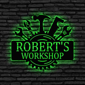 May include: Black metal workshop sign with green neon highlights. The sign displays tools such as a hammer, saw, and spanner, and the text "ROBERT'S WORKSHOP". It is mounted on a dark brick wall.