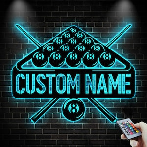 Op de afbeelding: Een zwart en blauw neonbord met een biljartrek met 8-ballen, gekruiste keuen en de tekst "CUSTOM NAME". Het bord is bevestigd aan een donkere bakstenen muur. Een afstandsbediening is rechtsonder zichtbaar.