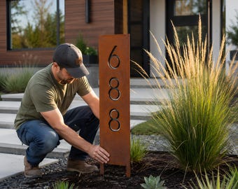 Puede incluir: Un letrero de dirección rectangular de metal marrón oxidado con los números 688 recortados. El letrero se está instalando en un parterre con mantillo oscuro y plantas decorativas. El letrero mide aproximadamente 122 cm de alto.