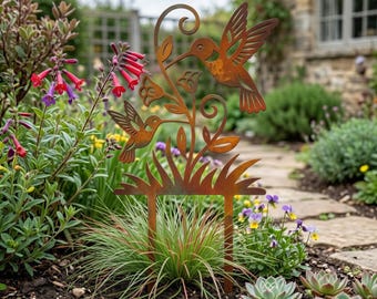 Può includere: Un paletto da giardino in metallo arrugginito con due colibrì, foglie e fiori. Il paletto è posto in un'aiuola con varie piante e fiori. La base del paletto è rettangolare.