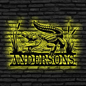 May include: A black metal alligator wall art piece with the name "ANDERSONS" in a yellow illuminated font. The alligator is detailed with open jaws, surrounded by cattails and grass, set against a dark brick wall.
