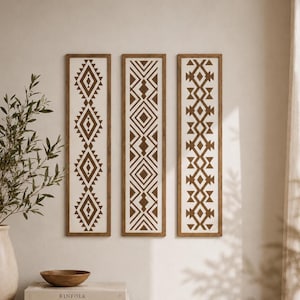 Op de afbeelding: Drie verticale rechthoekige wanddecoraties in houten lijsten, elk met een witte achtergrond en een opvallend bruin geometrisch patroon van ruiten, driehoeken en chevronlijnen in een herhalende verticale volgorde.