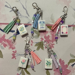 May include: Five keychains featuring white rectangular mahjong tiles with various floral and dragon designs, including one labeled Joker. Each keychain includes a colorful leather tassel, a string of pastel beads, and a silver metal lobster clasp.