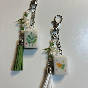 May include: Two keychains featuring white rectangular mahjong tiles with colorful floral and bird graphics. Each keychain includes a silver lobster clasp, a chain, white and green beads, a small flower charm, and a suede tassel in green or white.