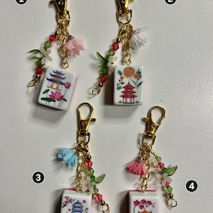 May include: Four gold-tone metal keychains featuring white rectangular mahjong tiles with colorful pagoda and floral illustrations. Each keychain includes a gold lobster clasp, a small fabric flower, and a strand of green and red beads.