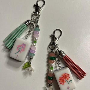 May include: Two keychains featuring white mahjong tiles with floral designs. One tile has a purple flower and the number 1, paired with a mint tassel and pastel beads. The other has a red flower and the number 4, with a coral tassel and pink and green beads.