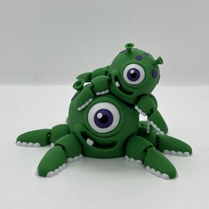 May include: A green, articulated monster figurine with a large eye and purple accents. A smaller monster is perched on top. The figurine has multiple jointed appendages and a smiling face. The design is whimsical and playful.