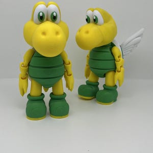 Puede incluir: Dos figuras de acción amarillas de Koopa Troopa. Las figuras tienen caparazones y botas verdes, con extremidades articuladas. Una figura tiene alas blancas. Las figuras tienen ojos verdes grandes y están sobre una superficie blanca.