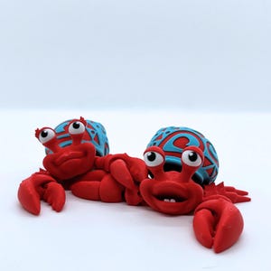 Puede incluir: Dos figuras de cangrejo rojas con rasgos de dibujos animados. Los cangrejos tienen grandes pinzas y caras expresivas. Sus caparazones son azules con un diseño rojo estampado. Las figuras están sobre un fondo blanco.