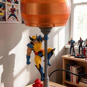 May include: A table lamp featuring a figure of the character Wolverine in a yellow and blue suit climbing a grey pole. The lamp has a round, orange, translucent shade and a base decorated with a small red fire hydrant and a textured surface.