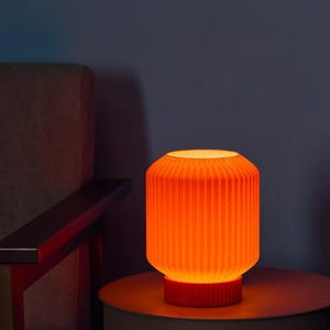 Könnte beinhalten: Eine zylindrische Tischleuchte mit einer leuchtend orangefarbenen, vertikal gerippten Außenseite. Die Lampe strahlt ein warmes Licht aus und verfügt über einen massiven, passenden orangefarbenen runden Sockel, der den strukturierten Schirm trägt.